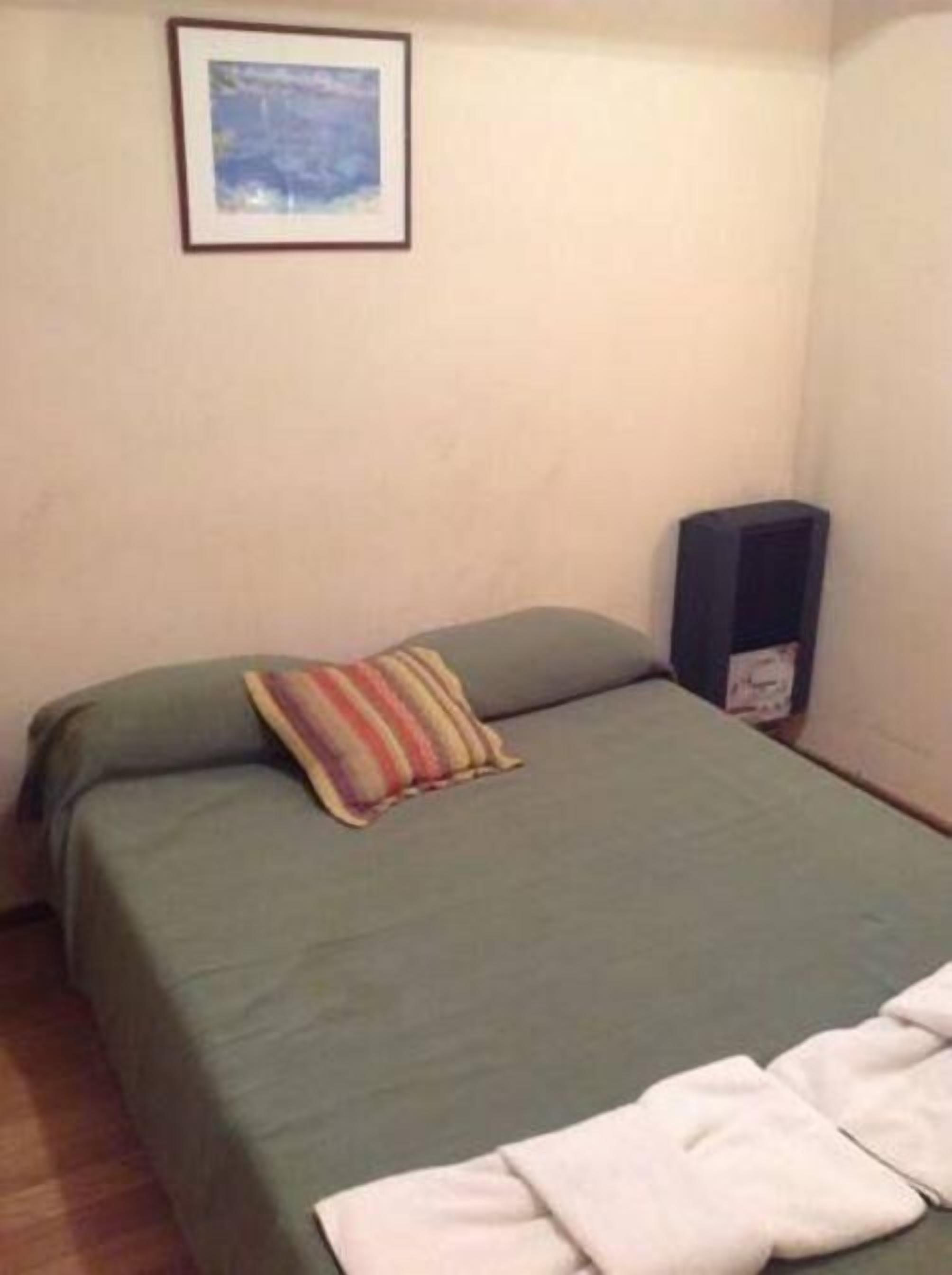 Una habitación pequeña y sencilla con una cama, un almohadón, un cuadro en la pared y un altavoz.