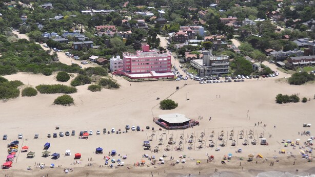 Vista aérea de una playa con un edificio grande, numerosos sombrillas y un helicóptero aterrizando en la arena.