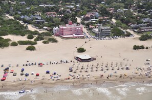 Vista aérea de uma praia com um edifício grande, vários guarda-sóis e um helicóptero pousando na areia.