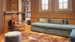 A spacious living room with a large sofa, a patterned ottoman, and a leather rug. There are two arched windows providing natural light, and a fireplace with a decorative mantel. - Ambit Boulevard Hotel Boutique (Santa Fe)