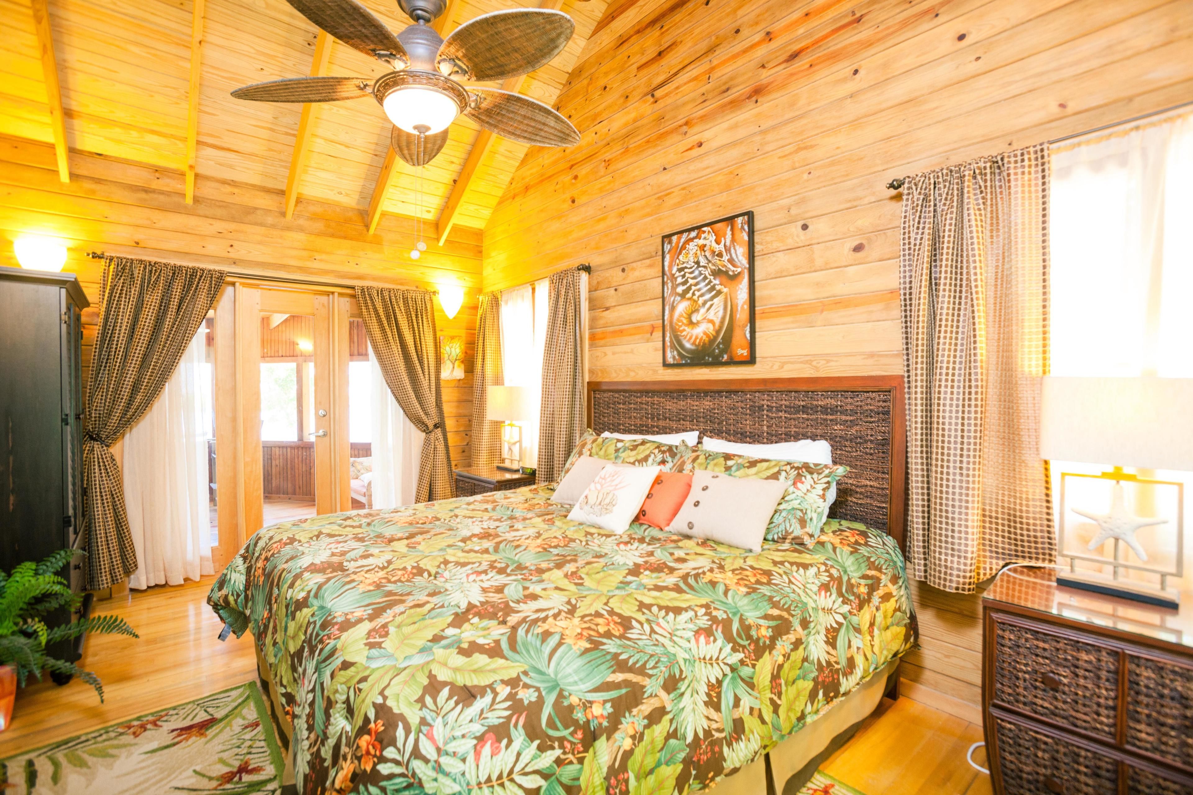 A wooden cabin room with a bed, ceiling fan, and a large window with curtains.