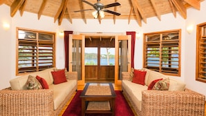 Una habitación acogedora con mobiliario de mimbre, ventilador de techo y ventanas grandes con contraventanas.