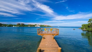 A wooden pier extending into a clear blue body of water with a wooden deck and two yellow chairs at the end. - Casa de Rexanna lower (Roatan)
