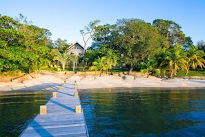 A wooden pier leading to a house with a white roof, surrounded by lush greenery and palm trees.