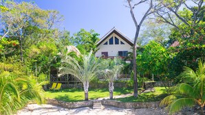 A house surrounded by lush greenery and a sandy area with a few palm trees. - Casa de Rexanna lower (Roatan)