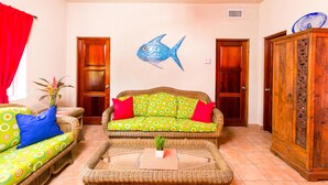 A living room with a green sofa, red cushions, a wicker coffee table, and a blue fish wall decoration. - Casa de Rexanna lower (Roatan)