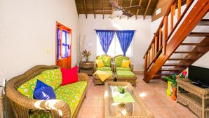 A living room with a sofa, two armchairs, a coffee table, and a TV. There is a wooden staircase and a window with blue curtains. - Casa de Rexanna upper (Roatan)