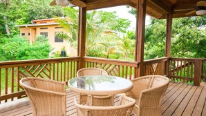 A wooden deck with wicker furniture and a glass table, overlooking a lush garden and a building. - Casa Feliz (Roatan)