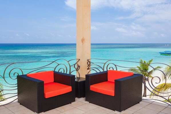Un balcón con dos sillas de mimbre y una mesa, con vistas al mar turquesa y un cielo despejado.