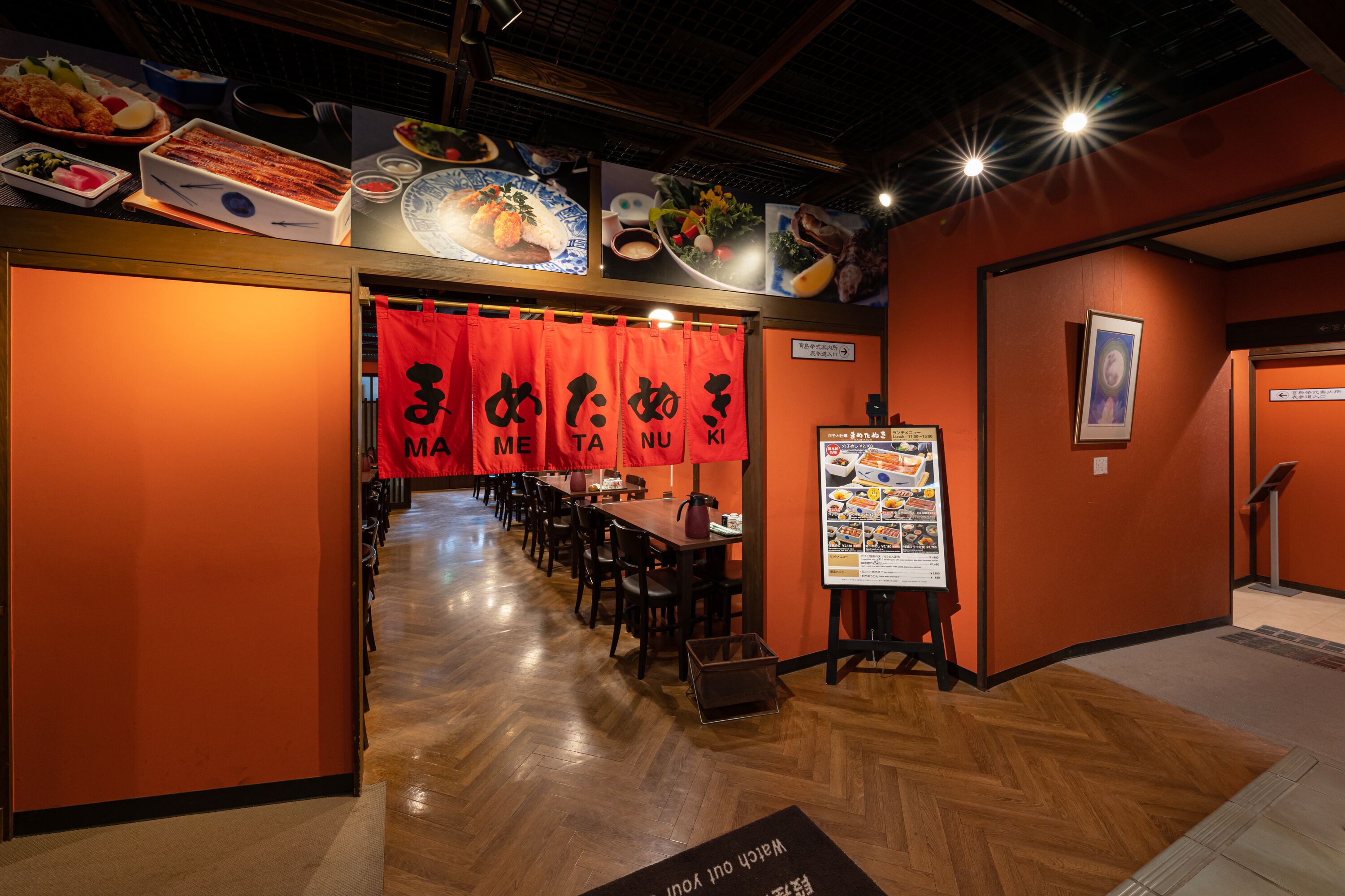 A restaurant interior with wooden floors, red banners, and a menu stand.