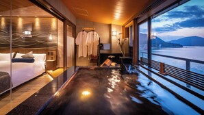 A modern hotel room with a large window overlooking a body of water, a bed, a seating area with a view, and a sauna. - Miyajima Kinsuikan (Hatsukaichi)