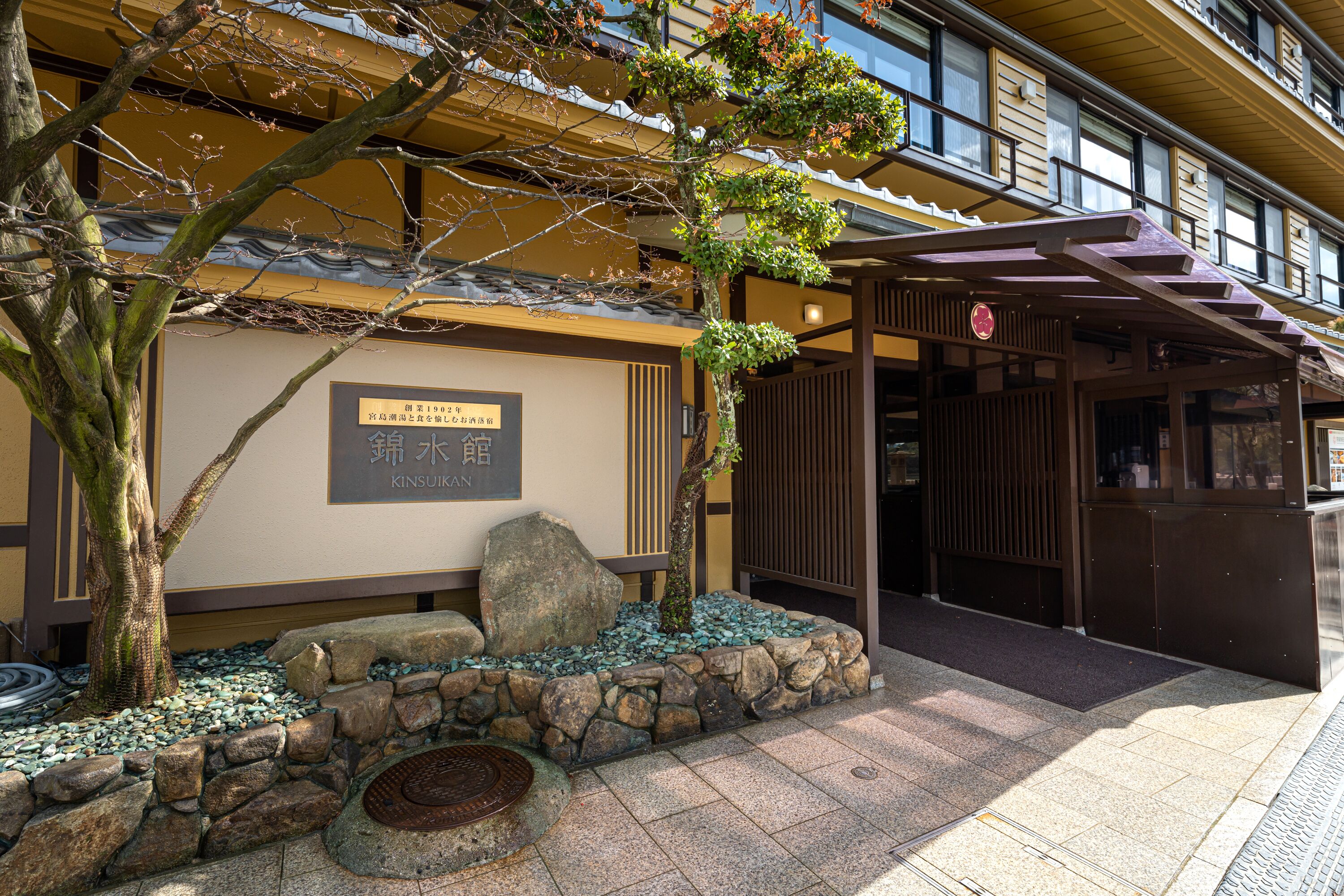 Een traditioneel Japans gebouw met een pad van stenen, een boom en een bordje met de tekst 'KINSUIKAN'.