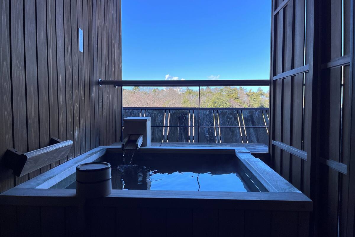Ein Holz-Wellnessbereich mit Whirlpool, einer Holzbank und Blick auf eine Holzdielen Terrasse und Bäume.