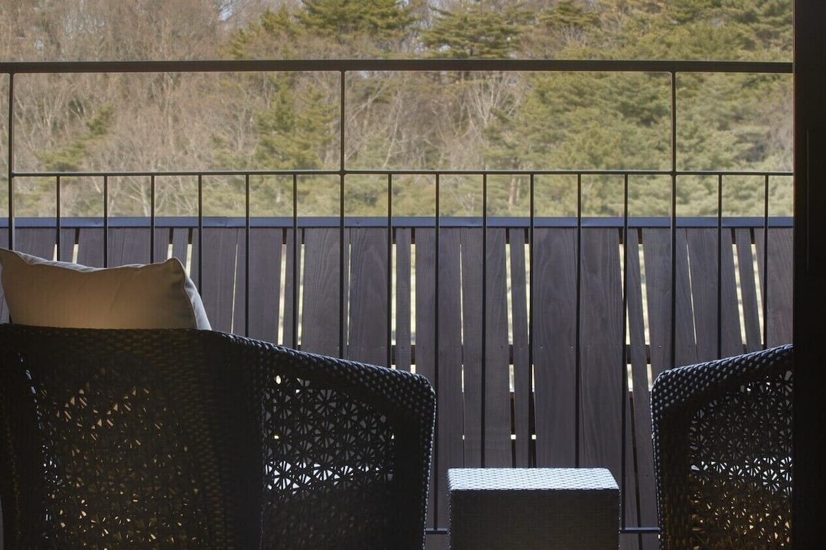 Ein Balkon mit zwei Korbstühlen und einem kleinen Tisch, mit Blick auf eine Holzterrasse und Bäume.