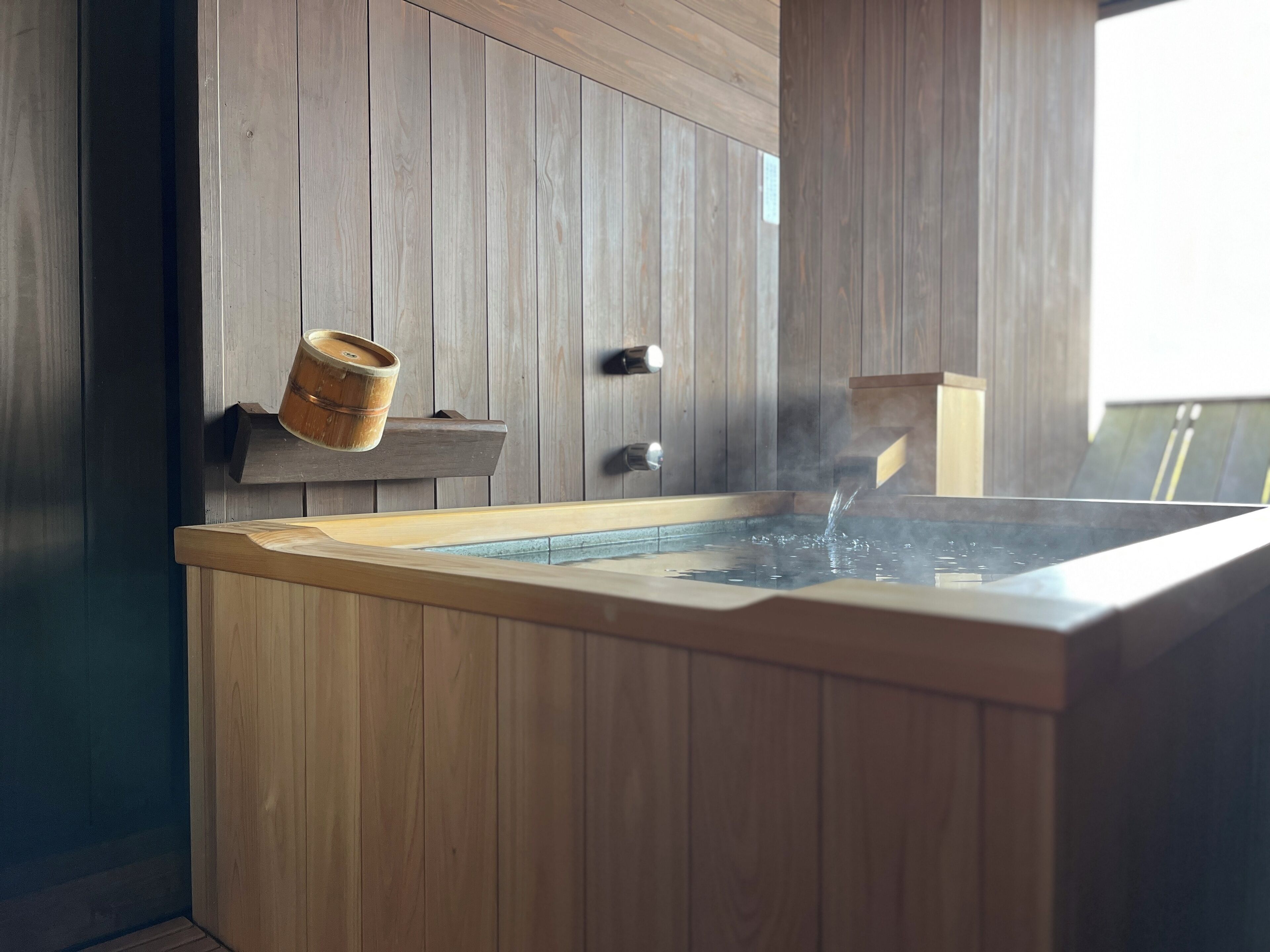 A wooden spa tub with water flowing into it, set against a wooden wall with a small wooden bucket on a shelf.