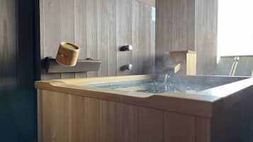 A wooden spa tub with water flowing into it, set against a wooden wall with a small wooden bucket on a shelf.