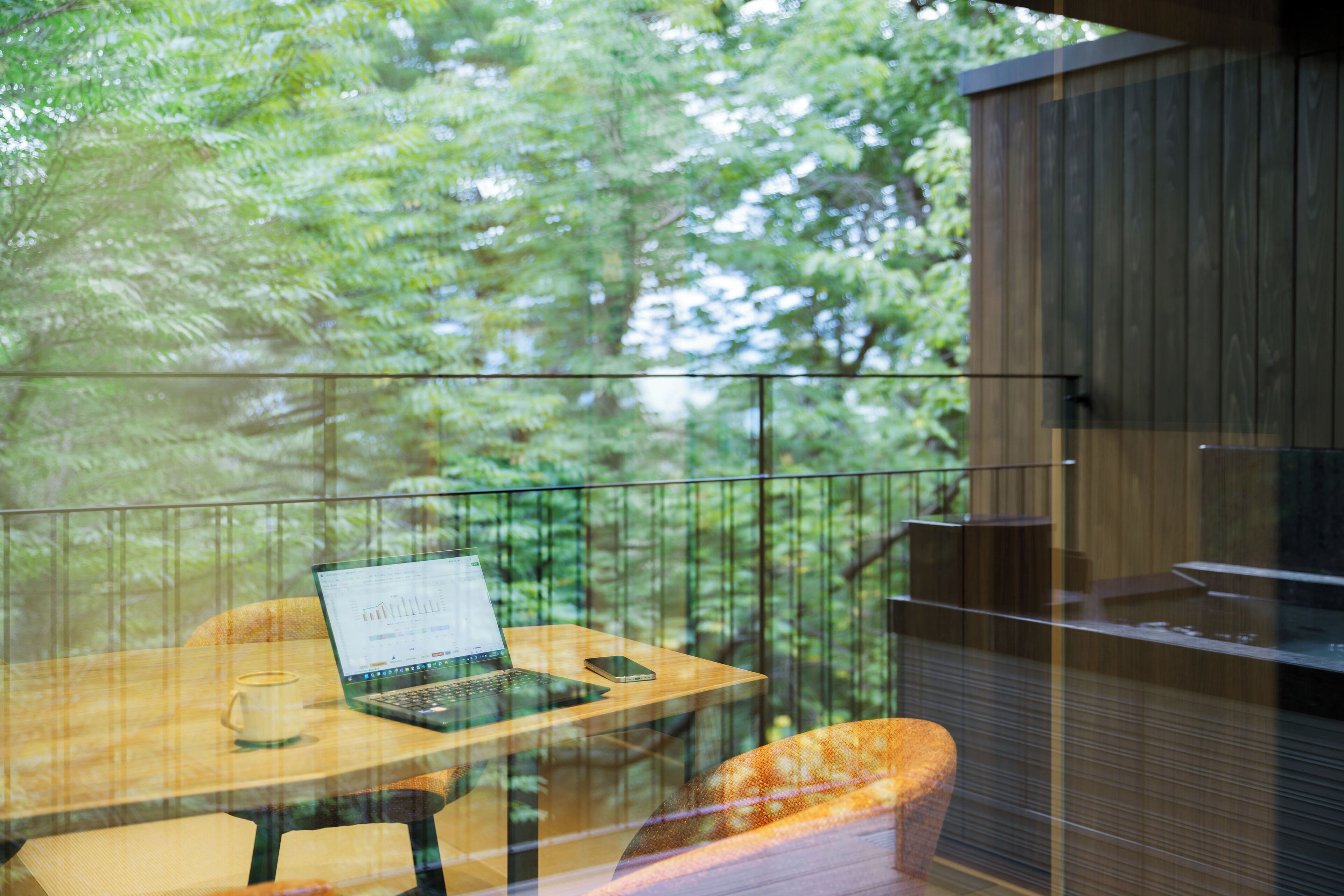 Un balcon vitré avec un ordinateur portable, un téléphone intelligent et une tasse posés sur une table en bois, donnant sur une zone boisée.