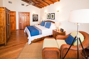 A bedroom with a wooden floor, a bed with blue and white bedding, a nightstand, a lamp, a wicker chair, and a ceiling fan.