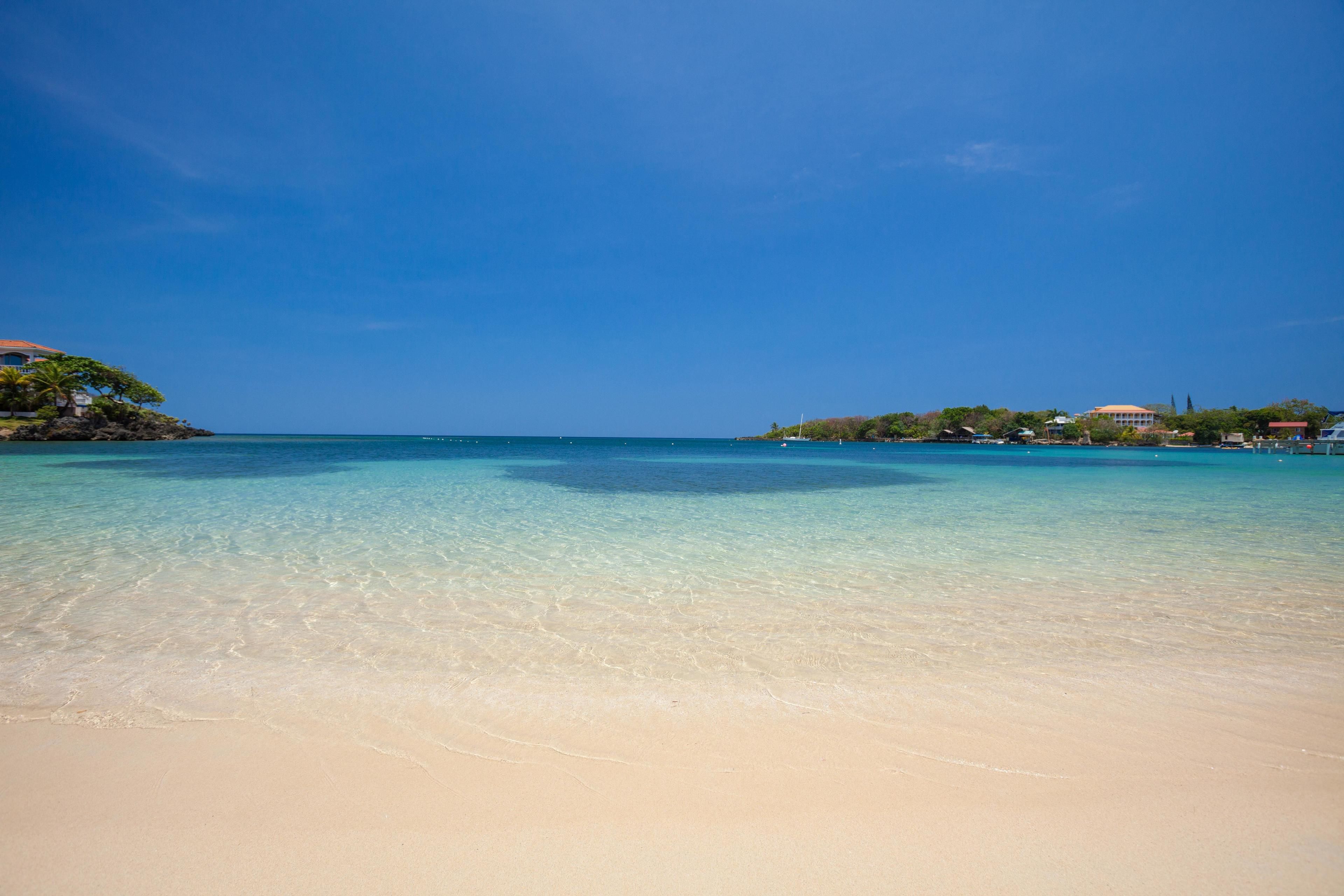 A beach with clear turquoise water, sandy shore, and buildings in the background.
