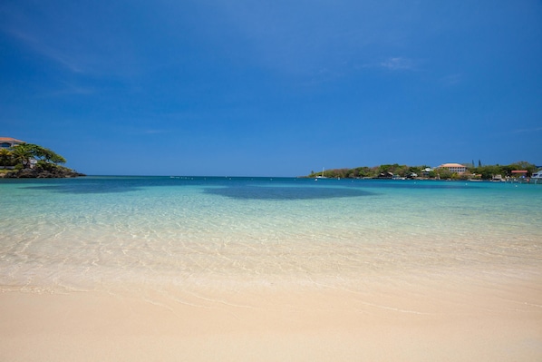 A beach with clear turquoise water, sandy shore, and buildings in the background.