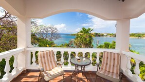 A covered balcony with two striped chairs and a small table, overlooking a scenic coastal view with clear blue water and greenery. - Coral Vista 5 3 Bedroom Option (Roatan)