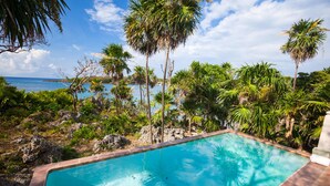 A rectangular pool with clear blue water, surrounded by lush greenery and palm trees, with a view of the ocean in the background. - Coral Vista 5 3 Bedroom Option (Roatan)