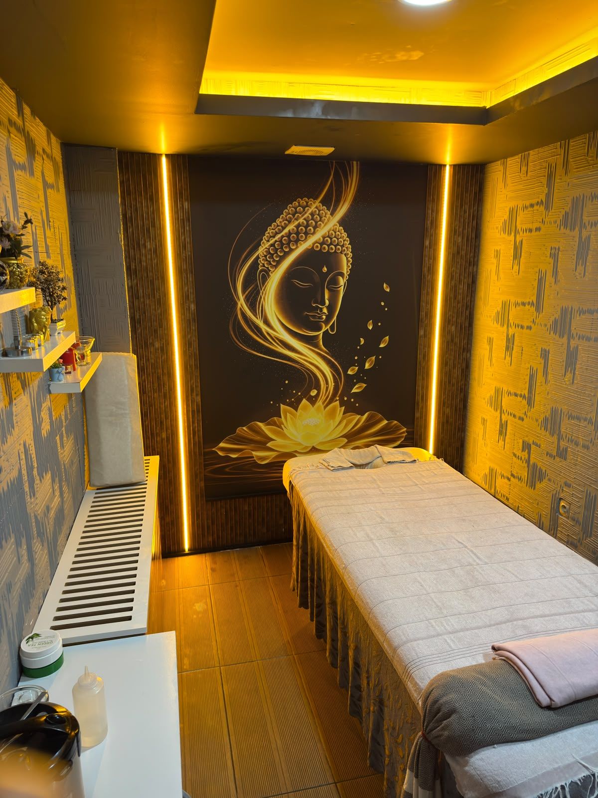 A room with a massage table, a wall with a Buddha and lotus design, and a small shelf with items.