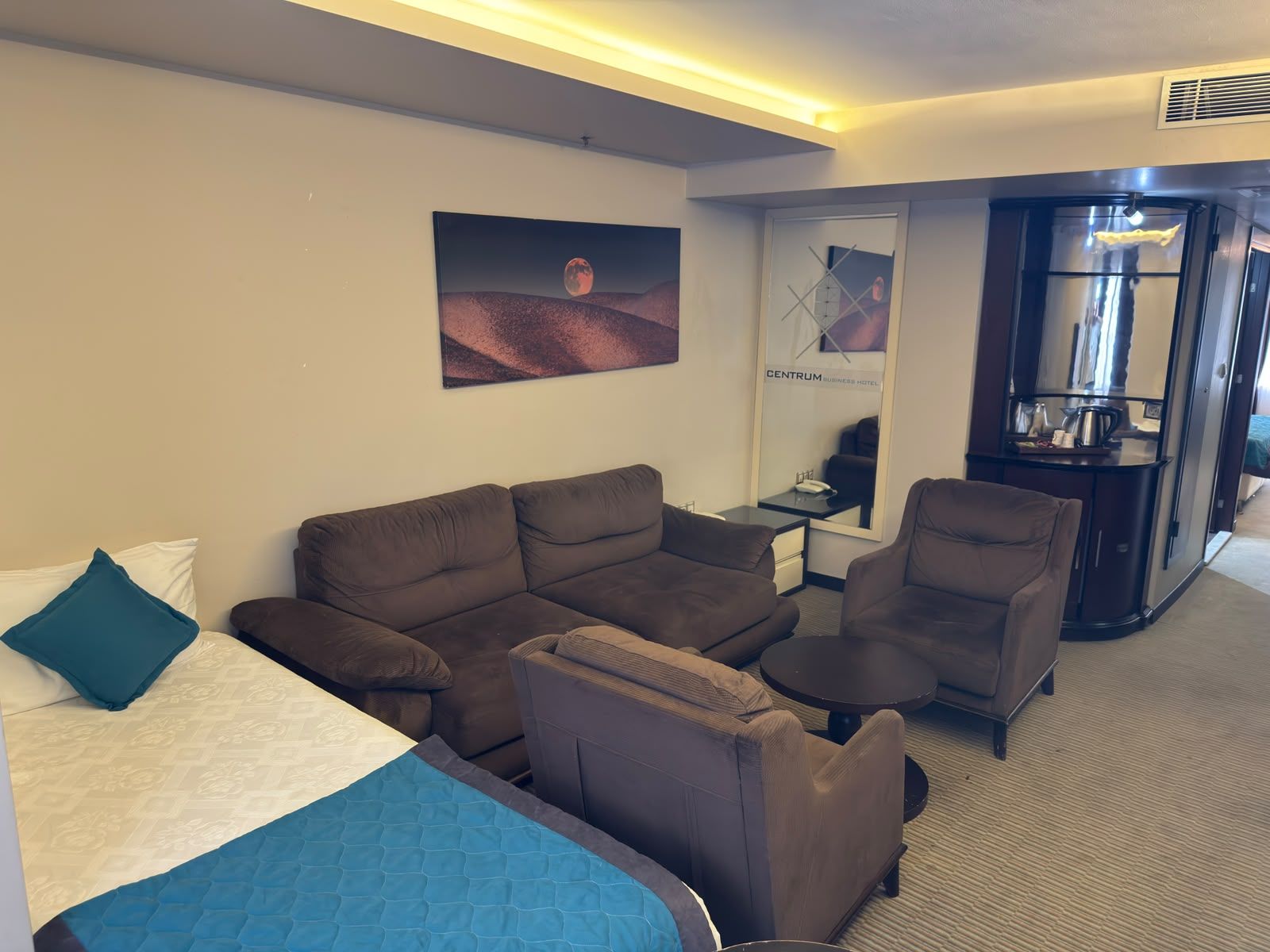 A hotel room with a bed, a sofa, a small table, a chair, a desk with a mirror, and a wall-mounted artwork.