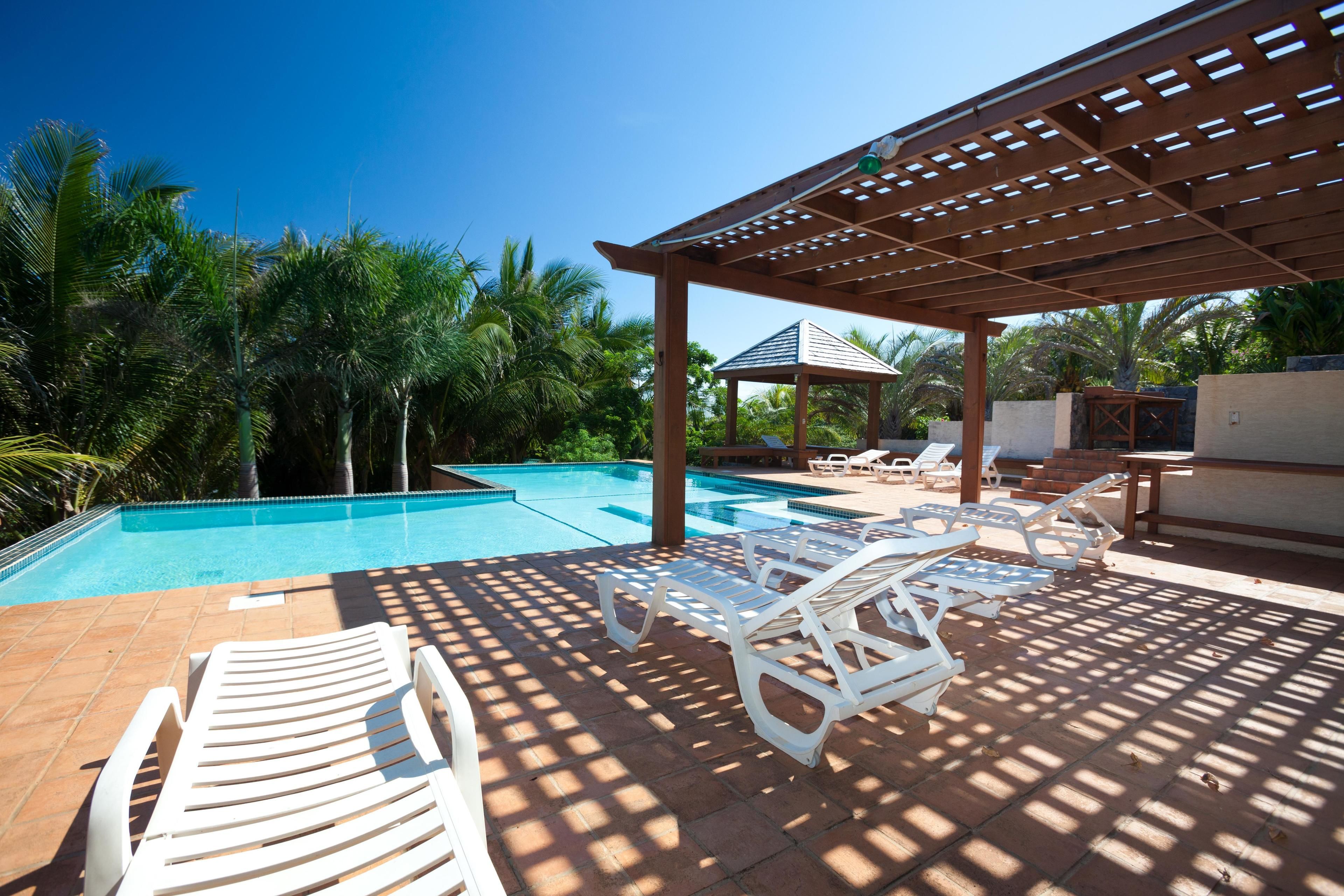 Un área de piscina con sillones blancos, una pérgola y un pequeño pabellón.