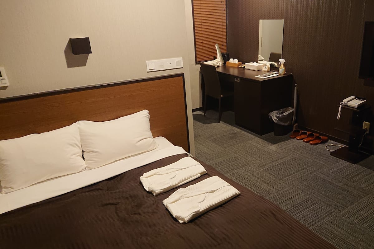 A hotel room with a bed, a desk, a mirror, a TV, and a trash bin.