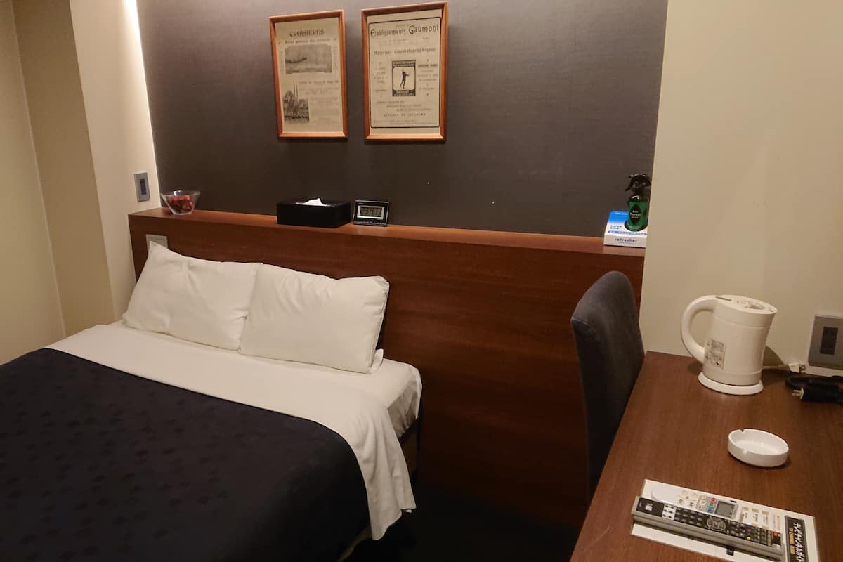 A hotel room with a bed, a wooden headboard, a nightstand with a clock, a coffee maker, and a framed certificate on the wall.