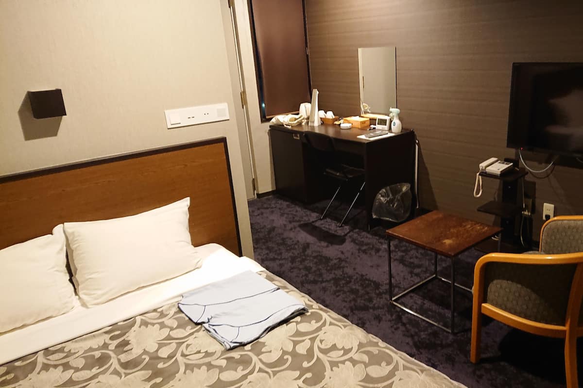 A hotel room with a bed, a desk, a chair, a TV, and a small table.