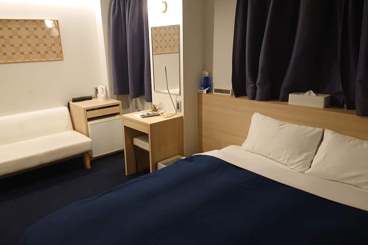 A hotel room with a bed, a desk, a chair, a small table, a nightstand, a mirror, and a window with curtains.