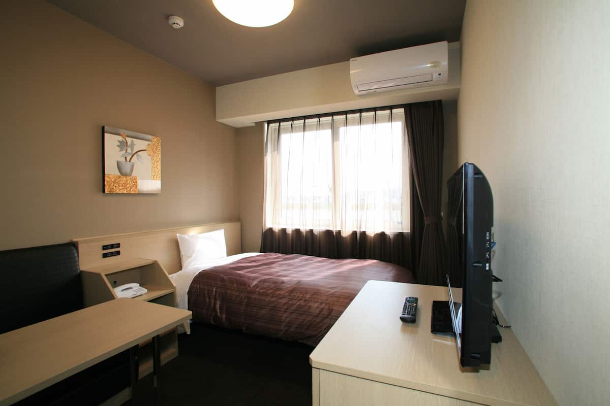 A hotel room with a bed, a desk, a chair, a television, and an air conditioning unit.
