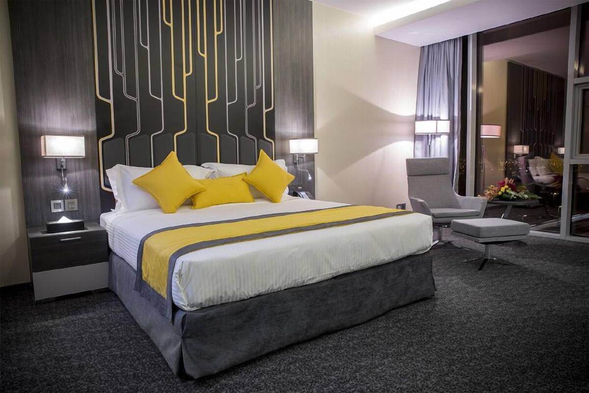 A modern hotel room with a large bed, two bedside tables with lamps, a grey upholstered armchair, and a small table with flowers.