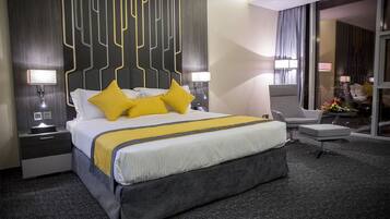 Een moderne hotelkamer met een groot bed, twee nachtkastjes met lampen, een grijze gestoffeerde fauteuil en een kleine tafel met bloemen.