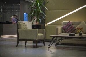 A lobby with a reception desk, a sofa with a cushion, a chair, a small table with a vase of flowers, and a large potted plant. - Executives Hotel Kafd (Riyadh)