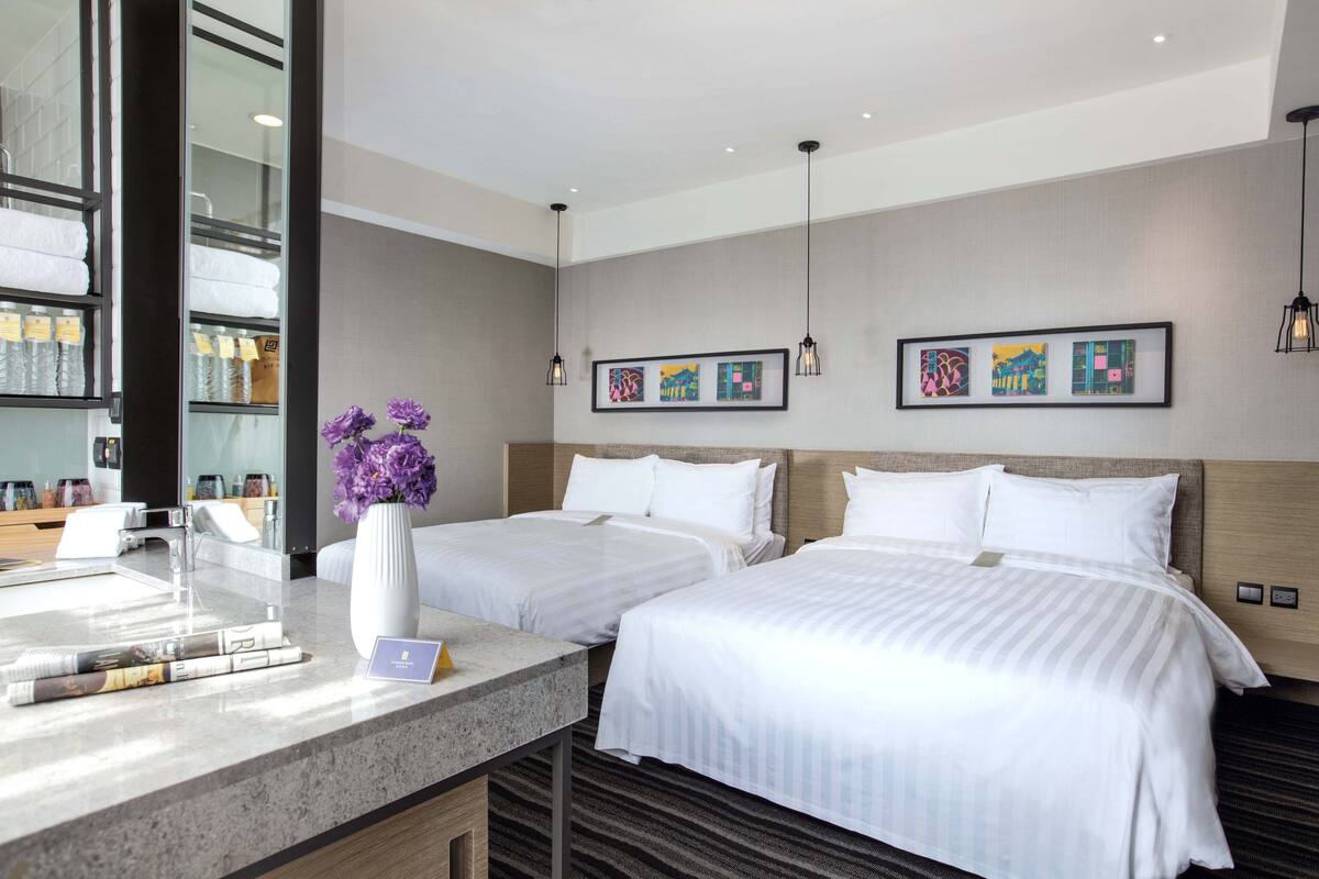 A hotel room with two beds, a vanity with a mirror, a sink, and a vase with flowers.