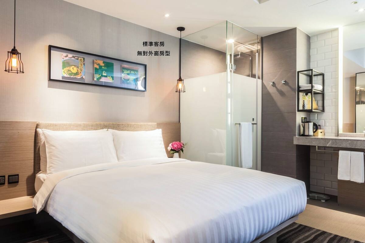 A hotel room with a bed, bedside controls, a wall-mounted picture, a shower area, and a bathroom with a sink and mirror.