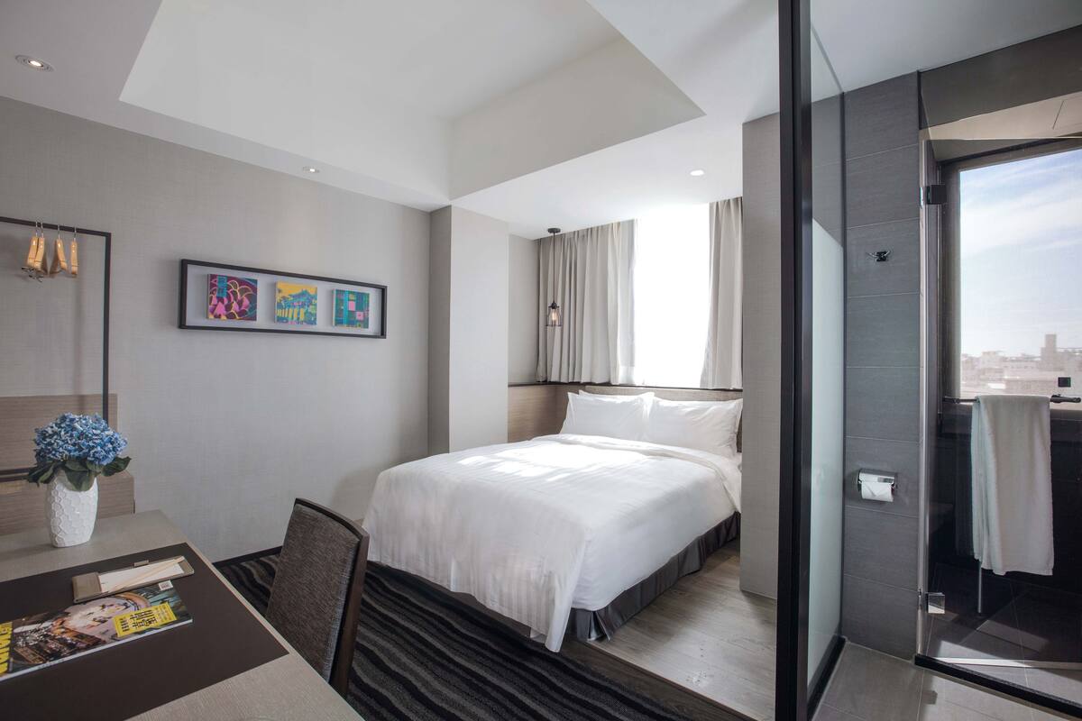 A modern hotel room with a large bed, a desk with a magazine, and a city view through the window.
