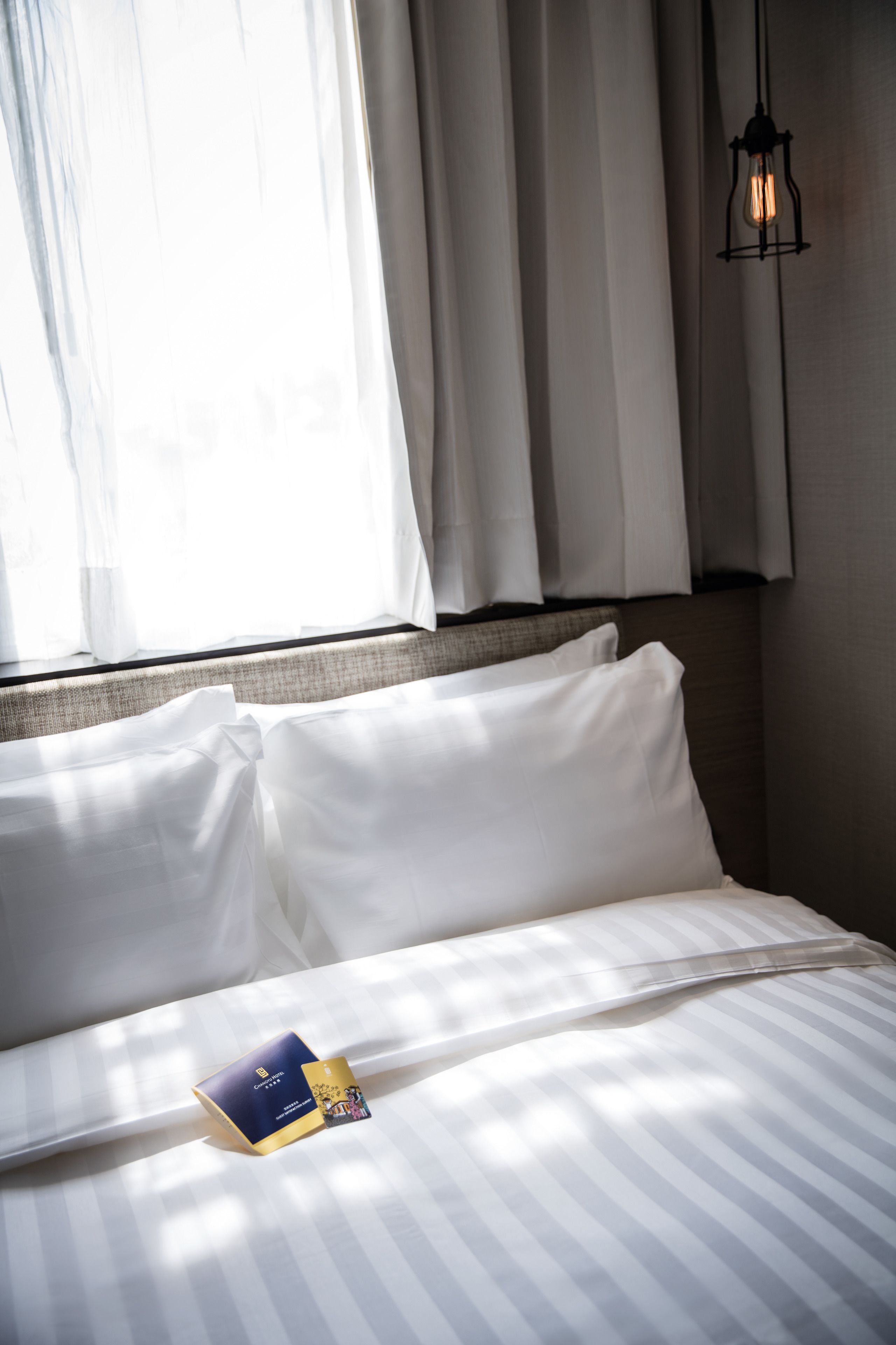 Un lit bien fait, avec du linge de lit blanc, une petite table de chevet sur laquelle repose un livre et une lampe suspendue au plafond.