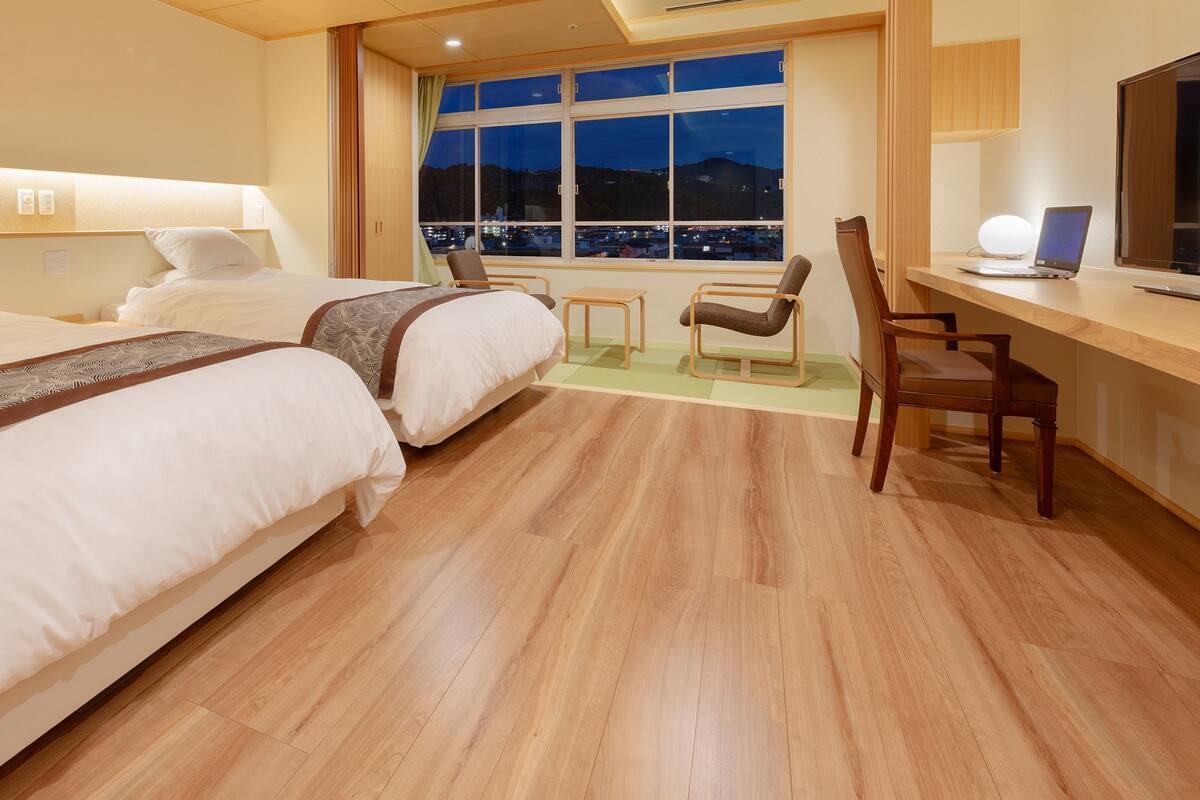 A hotel room with two beds, a desk, a chair, and a large window offering a city view.