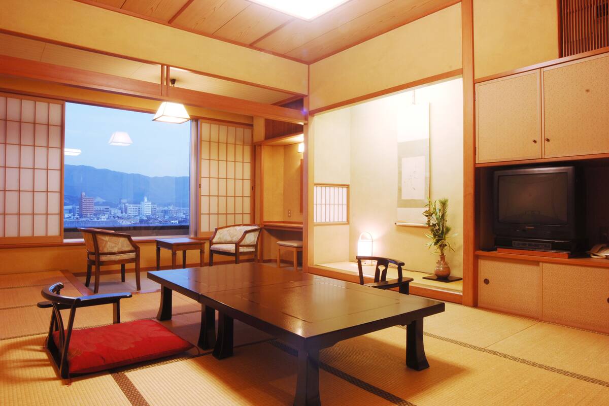 A traditional Japanese room with tatami flooring, a low table, chairs, a television, and a view of a cityscape through large windows.