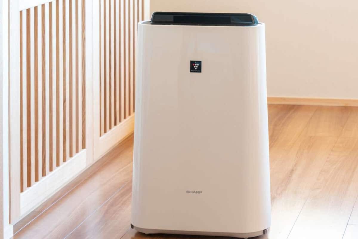 A white, modern air conditioning unit with a control panel on the top, placed on a wooden floor next to a white wall with vertical blinds.