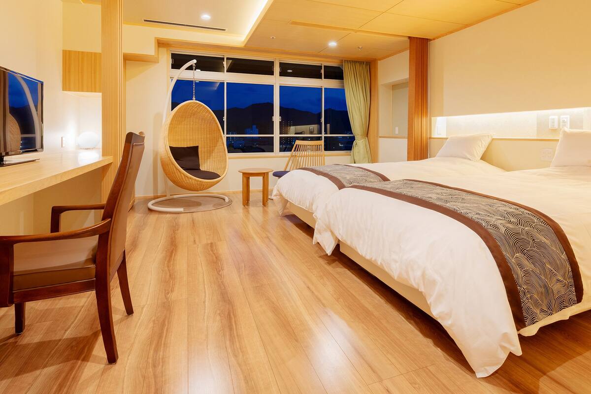 A modern hotel room with a wooden floor, a large bed with white bedding, a desk with a computer, a hanging chair, and a view of the city at night.