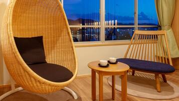 A room with a wicker chair, a small wooden table with two cups, and a view of a cityscape at dusk.