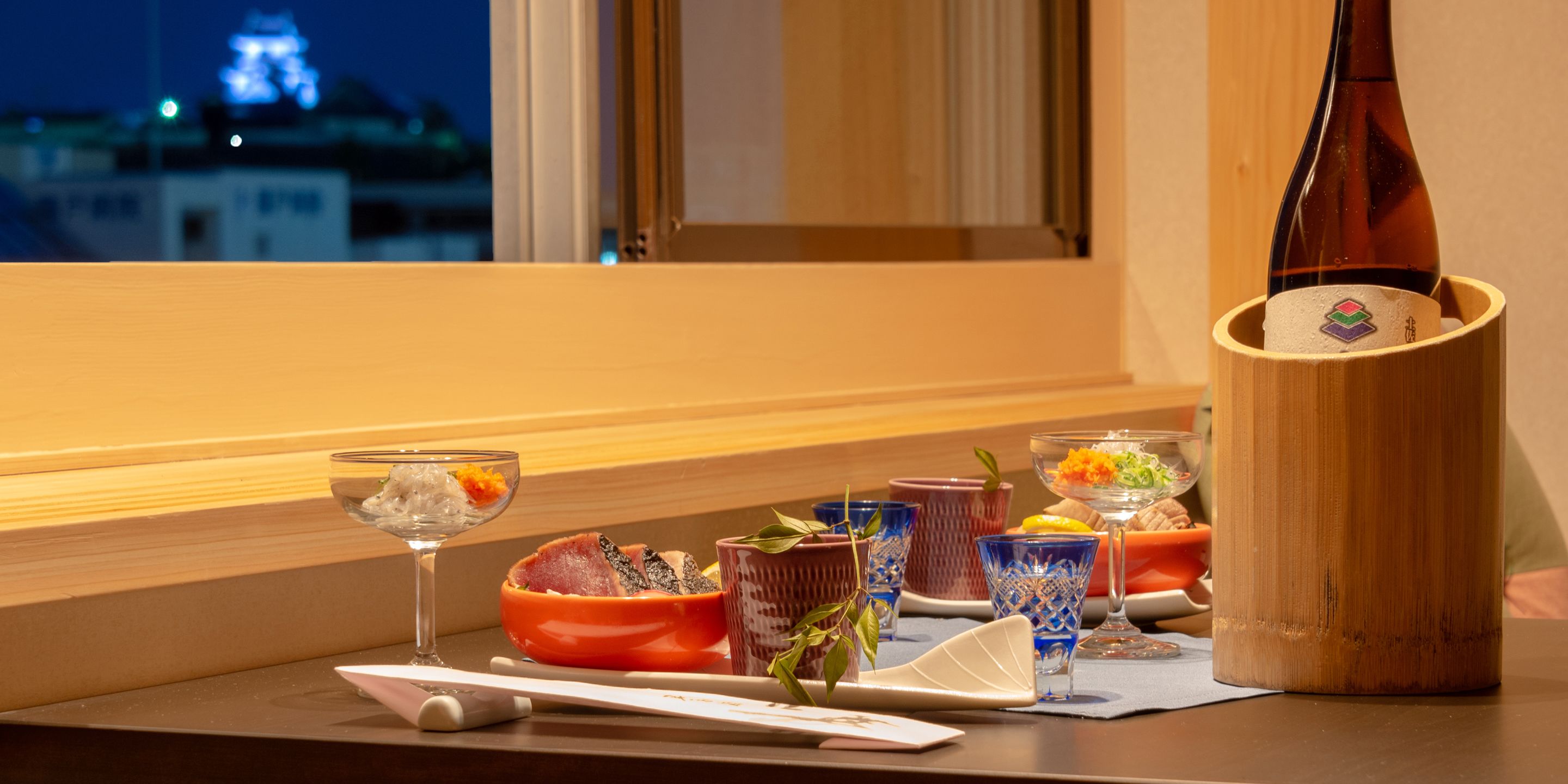 A table with a bottle, glasses, and a bowl of food, with a window view in the background.