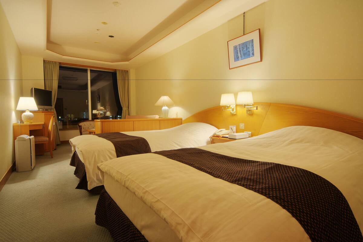 A hotel room with two beds, a desk with a TV, and a window with curtains.