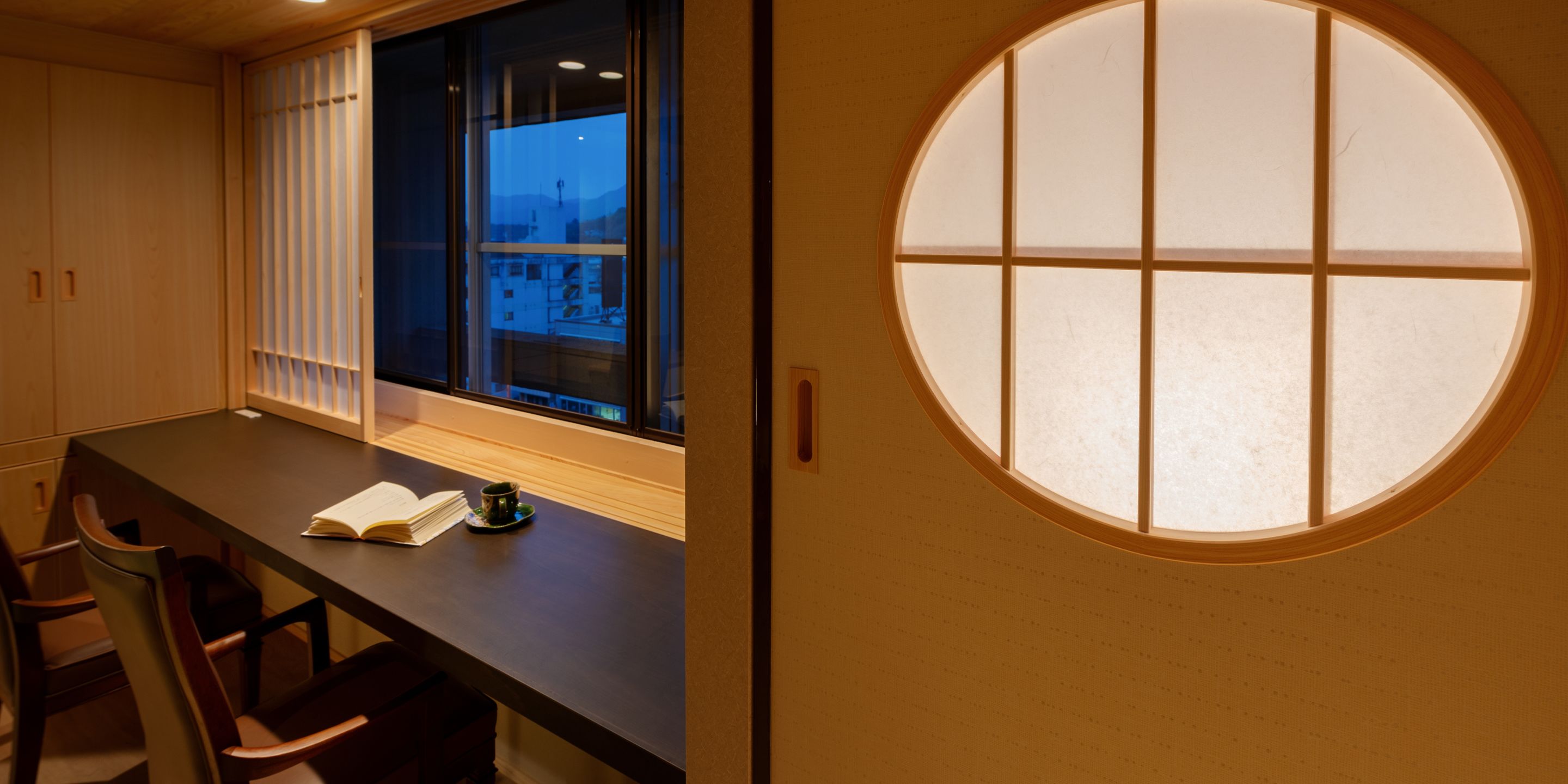 A room with a wooden desk, a book, and a cup. There is a window with blinds and a round window on the wall.