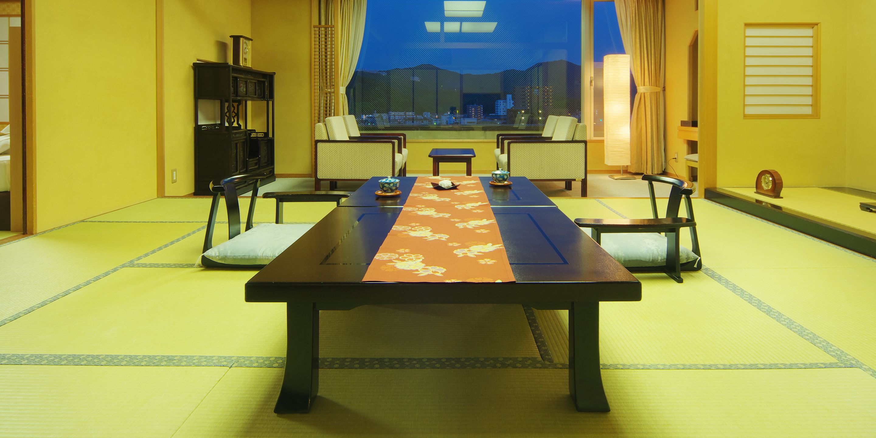 A traditional Japanese-style room with tatami flooring, a low table with a table runner, and a view of the cityscape through large windows.